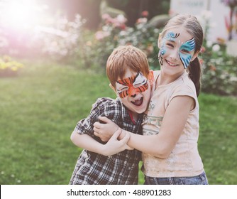 Happy Children With Face Art Paint In Park. Boy And Girl Outdoors On Child Birthday Masquerade Party Have Fun, Laugh And Hug Each Other As Friends Or Siblings. Entertainment And Holidays.