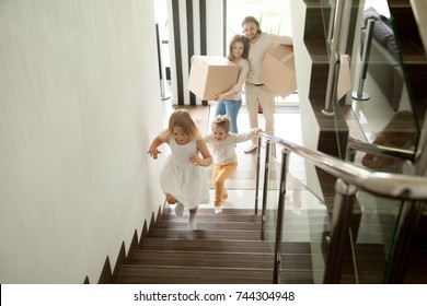 Happy Children Going Upstairs Inside Two Story Big House, Excited Kids Having Fun Stepping Walking Up Stairs Running To Their Rooms While Parents Holding Boxes, Family Moving In Relocating New Home 