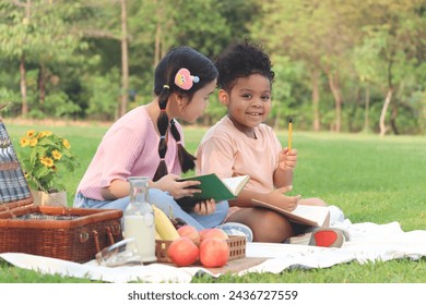 Happy children have a picnic in summer park, cute curly hair African girl with Asian buddy friend studying, reading book together while sitting on mat on green grass. Kid do homework in outdoor garden - Powered by Shutterstock