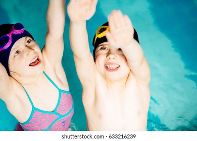 Happy Children Holding Hands Up And Waving In Swim Pool, View From Above