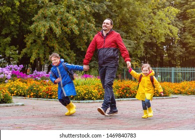 Happy Children Laugh, Rush And Run To School, Dressed In Raincoats, With A Briefcase Behind A Backpack. Dad Leads His Daughter And Son Daroga To School In Rainy Weather.close-knit Family Holding Hands