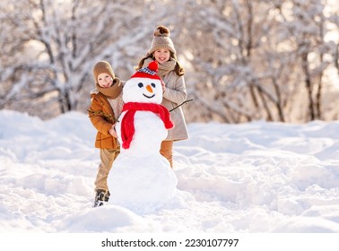 Happy children little brother and sister dressed in warm stylish clothes standing in winter snowy park broadly smiling while proudly posing near snowman made by them, kids playing with snow outdoors