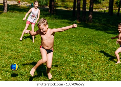 Happy Children Playing Dodgeball With The Ball On The Green Lawn In The Summer