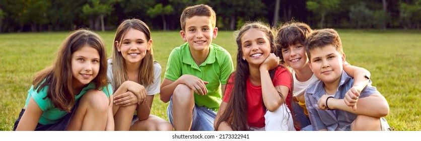 Happy Children Sitting On Green Grass And Smiling. Cheerful Little Kids Enjoying Their School Break, Spending Time In A Summer Camp, Making New Friends, And Playing Outside. Group Portrait. Banner