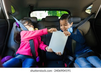 Happy children smiling and playing with a tablet while sitting in the back seat while going on a road trip drive - Powered by Shutterstock