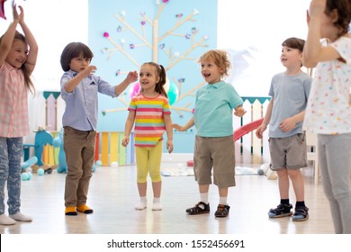 Happy Children Stand Semicircle On Floor In Kindergarten Or Day Care Centre. Preschool Kids Have Fun Indoors, Playing Games