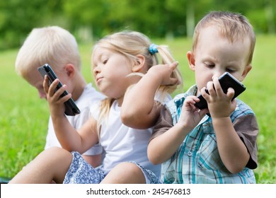 Happy Children Using Smartphones Sitting On The Grass. Brothers And Sister.