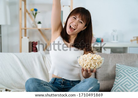 happy chinese lady raising her hand is excited watching the olympic games in the living room. beautiful asian female shouts with glee for the team she supports wins championship.
