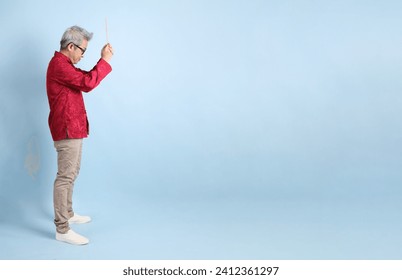 Happy Chinese new year. Asian Chinese energetic senior man wearing red traditional cheongsam qipao or changshan dress with gesture of hand holding the incense sticks  isolated on blue background. - Powered by Shutterstock