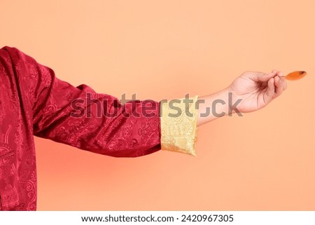 Happy Chinese new year. Hand of asian chinese senior man wearing red traditional clothing with gesture of hand holding golden spoon isolated on orange background.