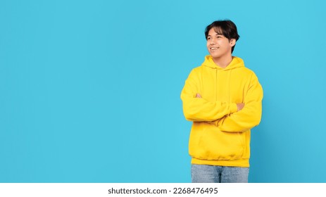 Happy Chinese Teen Boy Looking Aside At Free Space For Text Standing Crossing Hands Over Blue Studio Background, Wearing Yellow Hoodie. Male Teenager Posing Advertising Great Offer. Panorama - Powered by Shutterstock