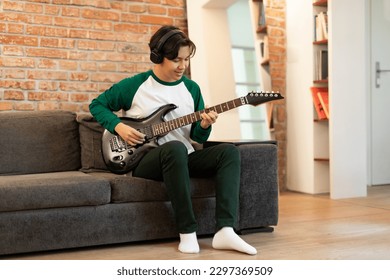 Happy Chinese Teenager Guy Learning Music Playing Electric Guitar And Wearing Earphones Indoors. Talented Musician Boy Posing With Musical Instrument Sitting On Sofa At Home. Hobby And Leisure - Powered by Shutterstock