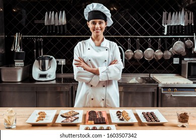 Happy Chocolatier In Chef Hat Standing With Crossed Arms Near Tasty Chocolate Candies On Plates