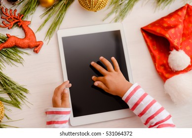 Happy Christmas background. Top view on child hands using tablet computer with toys and Santa Claus hat on light wooden table. Modern festive decoration ready for joyful message or shopping time - Powered by Shutterstock