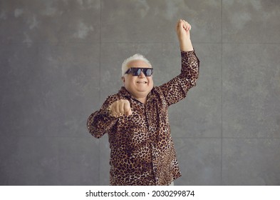 Happy Chubby Senior Man Vibing To Music. Portrait Of Funny Cheerful Rich White Haired Fat Old Guy In Cool Glasses And Leopard Patterned Party Shirt Dancing And Having Fun On Grey Studio Background