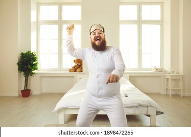 Happy Chubby Young Man Wakes Up One Weekend Morning, Gets Up From Bed And Enjoys Free Time At Home. Funny Cheerful Confident Fat Guy In White Pajamas Dancing Gangnam Style In Light Sunny Bedroom