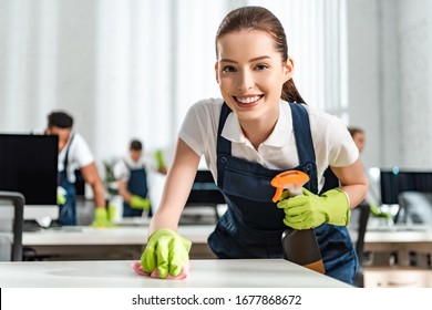 happy cleaner looking at camera while cleaning office desk with rag