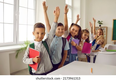 Happy clever school children ready for class. Small group of cheerful cute smart kids in casual clothes with books and backpacks standing near row of desks in classroom, raising up hands and smiling - Powered by Shutterstock