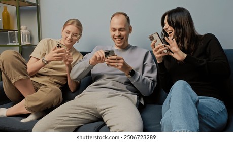 Happy close friends enjoying time together playing with their phones in cozy setting. Deeply immersed in digital world. Excited expression sitting on sofa. Modern social interactions concept - Powered by Shutterstock