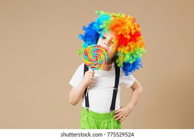 Happy clown boy in large colorful wig. Let's party! Funny kid clown. 1 April Fool's day concept. Portrait of a child eating lollipop. Birthday boy. Brazilian Carnival. Venice Carnival.