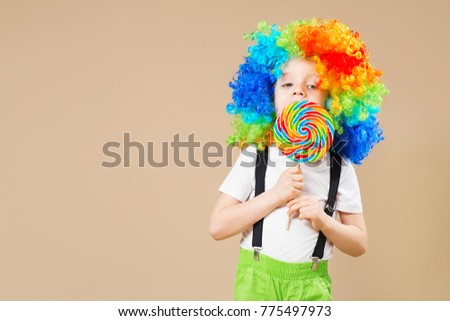 Happy clown boy in large colorful wig. Let's party! Funny kid clown. 1 April Fool's day concept. Portrait of a child eating big lollipop. Birthday boy.