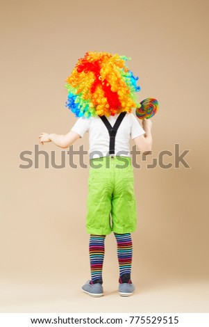 Happy clown boy in large colorful wig. Let's party! Funny kid clown. 1 April Fool's day concept. child eating lollipop. Birthday boy. Back view