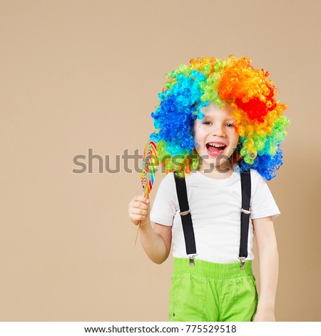 Happy clown boy in large colorful wig. Let's party! Funny kid clown. 1 April Fool's day concept. Portrait of a child eating lollipop. Birthday boy.