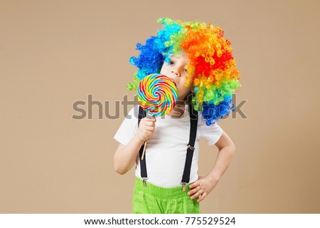 Happy clown boy in large colorful wig. Let's party! Funny kid clown. 1 April Fool's day concept. Portrait of a child eating lollipop. Birthday boy. Brazilian Carnival. Venice Carnival.
