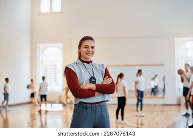 Happy coach standing with her arms crossed at school gym and looking at camera. The students are int he background. - Powered by Shutterstock