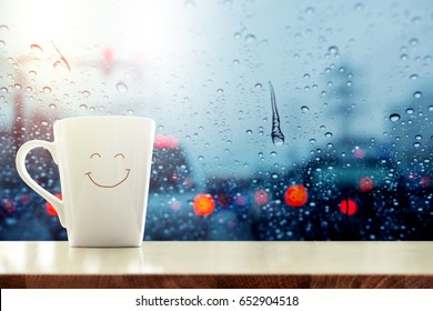 Happy Coffee Mug with smiley face on desk inside glass window, Blurred traffic jam light in city as outside view, Relaxing in cafe or Rest on rainy day