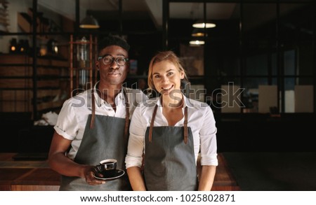 Happy coffee shop owner couple standing inside their shop. Man and woman baristas standing inside their cafÃ?Â© wearing apron with the man holding coffee cup.