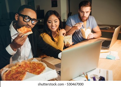 Happy colleagues working late at night with laptops while eating pizza. - Powered by Shutterstock