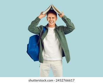 Happy college or high school student having fun with book. Studio portrait of funny cheerful boy with backpack standing isolated on light background holding textbook on top of his head and laughing - Powered by Shutterstock