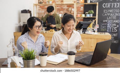 Happy College Student Girls Celebrate Successful Teamwork Results In Cafe Bar. Excited Two Asian Woman Working On Project And Pass Exam In Coffee Shop. Startup In Coffeehouse Male Owner In Counter