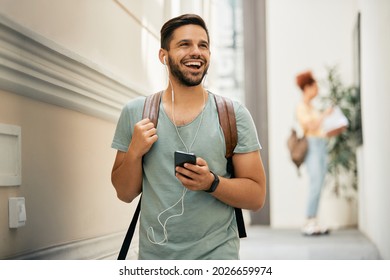 Happy College Student Using Mobile Phone And Listening Music Over Earphones While Walking Down The Hall. 