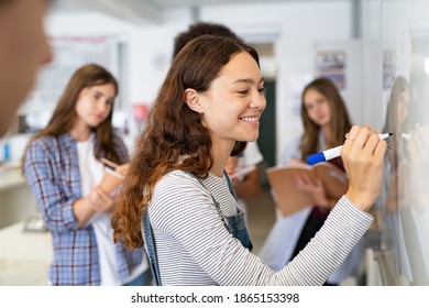 Happy college student writing equation on white board in class. Satisfied young girl solving math problem on whiteboard with classmates in background watching her. Proud high school student writing.