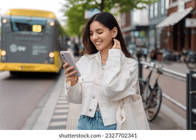 Happy commute on public transport. Multi-ethnic attractive woman with beautiful smile at bus station looking at phone - Powered by Shutterstock