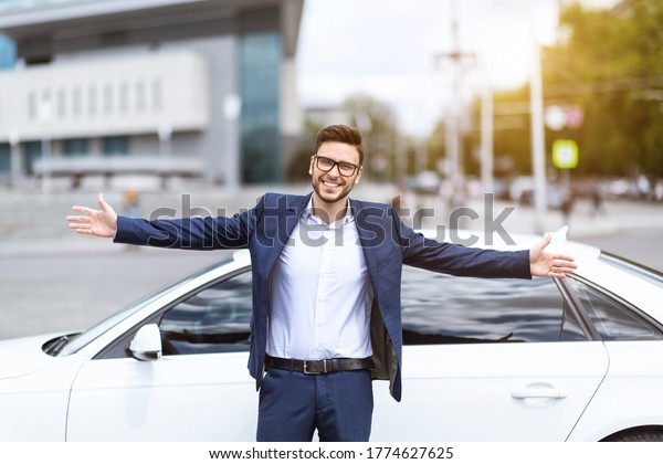 Happy company leader standing with open arms in\
front of his car at city\
street