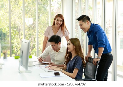 Happy Concentration At Meeting. Group Of Young Business People Working And Communicating While Sitting And Standing In Office Desk Together With Computer Laptop And Office Supply In Window Background