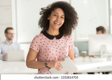 Happy confident african american business woman employee holding digital tablet looking at camera standing in office, smiling millennial mixed race female intern manager young professional portrait