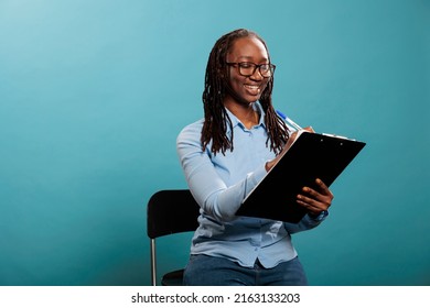 Happy Confident African American Woman With Clipboard Documentation Signing Agreement Papers On Blue Background. Attractive Young Adult Person Writing Down Names On Checklist. Studio Shot