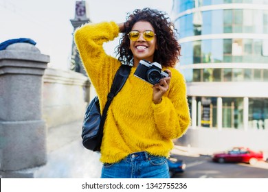 Happy Confident  African Woman Holding Photo Camera And Walking In Big Modern City. Wearing Trendy Spring Outfit And Back Pack. Traveling Black Female.