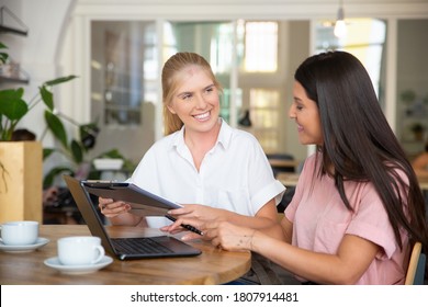 Happy Confident Agent And Customer Meeting Over Cup Of Coffee And Signing Agreement. Young Women Sitting At Table With Open Laptop And Analyzing Document. Consulting Or Cooperation Concept