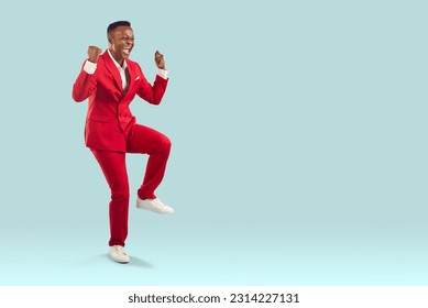 Happy confident attractive African American man in cool modern trendy red suit celebrating victory, feeling overjoyed and super excited, fist pumping and having fun on light blue copyspace background