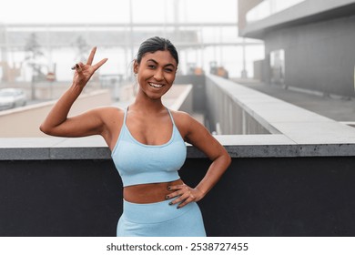 Happy confident fitness model gesturing peace sign while standing on rooftop on blurred city background, enjoying healthy lifestyle and urban training - Powered by Shutterstock