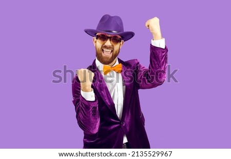 Happy confident man celebrating success. Funny excited cheerful bearded guy in hat, suit and orange bow tie standing on purple background, fist pumping and shouting Yes, Yay, I did it, Cool, Hurray