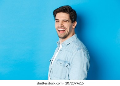 Happy And Confident Man Turn Head At Camera, Winking And Smiling, Standing Over Blue Background