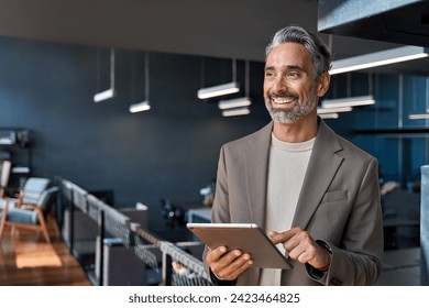 Happy confident mature business man executive standing in corporate office using digital tablet. Smiling middle aged professional businessman manager wearing suit looking away at work in modern space. - Powered by Shutterstock