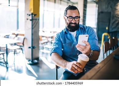 Happy Confident Mature Male Entrepreneur In Casual Wear Sitting Near Bar Station In Cafe, Cheerful Hospitable Bearded Man Owner Of Small Business Coffee Shop Checking Notification On Smartphone