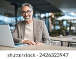 Happy confident mature professional business man hybrid working looking at camera. Smiling middle aged businessman wearing glasses sitting at outdoor office table using laptop. Portrait. Copy space.